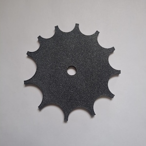 May include: A black, circular gear-shaped component with a textured surface and twelve pointed teeth along the outer edge. The object features a single, centered circular hole, providing a flat, uniform appearance suitable for mechanical assembly.