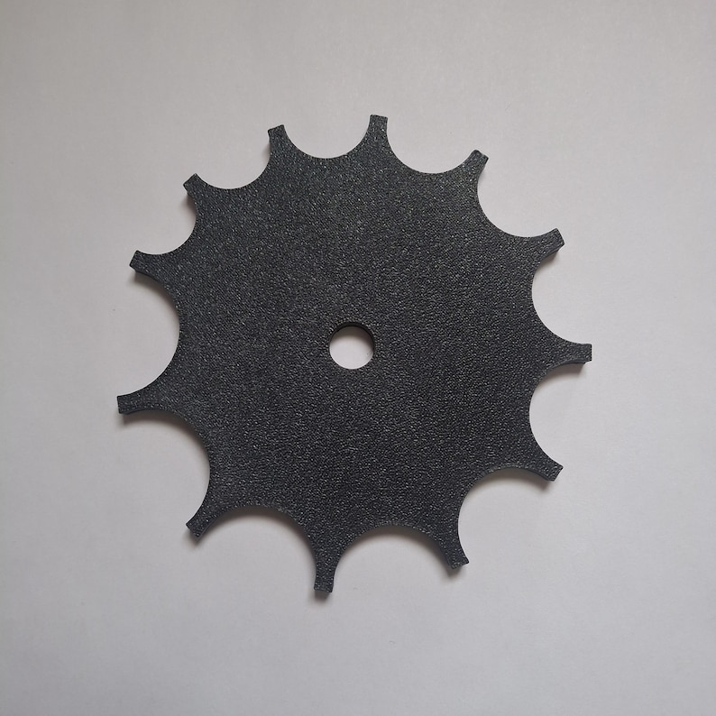 May include: A black, circular gear-shaped component with a textured surface and twelve pointed teeth along the outer edge. The object features a single, centered circular hole, providing a flat, uniform appearance suitable for mechanical assembly.