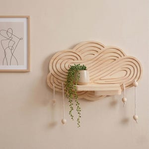 Op de afbeelding: Een houten, wolkvormig rek met een klein plankje en hangende houten kralen. Een kleine beige pot met een hangende groene plant staat op het rek. Een ingelijste lijntekening hangt aan de muur.