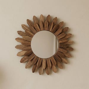 May include: A round decorative mirror with a wooden frame. The frame is made of individual wooden pieces arranged in a sunburst pattern. The mirror has a light brown color and is mounted on a neutral-colored wall.