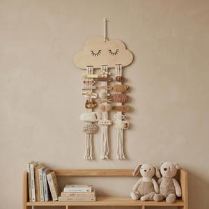 Puede incluir: Un soporte de pinzas para el pelo en forma de nube de madera con detalles de macramé. La nube tiene pestañas dibujadas y contiene varias pinzas decorativas en tonos beige, marrón y rosa. El soporte cuelga de una pared beige sobre una estantería de madera.