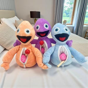 Op de afbeelding: Drie pluche speelgoed in oranje, paars en blauw, elk met een lachend gezicht, grote ogen en een rits aan de voorkant. Elk speeltje heeft een rode bal en een roze hartvormig object aan de binnenkant. Het speelgoed ligt op een wit bed.