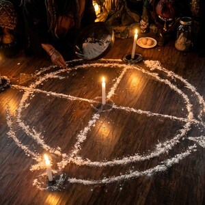May include: A ritual scene with a circle drawn on a wooden floor, illuminated by lit candles. People are gathered around the circle, some holding bowls. The scene is dimly lit, creating a mysterious atmosphere. The circle is made of a white powder.