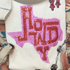 May include: A cream-colored t-shirt with a pink glitter Texas outline and the word "HOWDY" in red glitter. The image includes a cowboy hat, white boots, earrings, and a decorative skull.