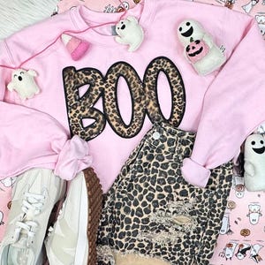 May include: A pink sweatshirt with the word "BOO" in leopard print, paired with leopard print denim shorts and white sneakers. The image includes Halloween-themed plush ghost decorations, creating a festive, casual outfit.