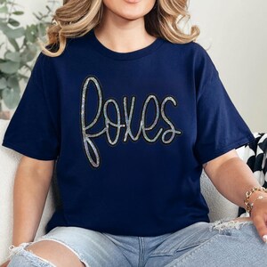 Product Foxes Sequin Patch, a sparkling sequin embroidered patch of the word "foxes" on a navy blue t-shirt.