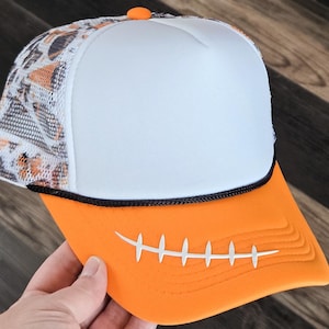 May include: A trucker hat with a white front panel, an orange bill, and a mesh back with a football pattern. The orange bill features white stitching, and the hat has a black trim and an orange button on top.