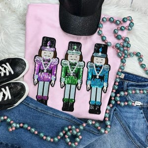 May include: A pink sweatshirt with three sequined nutcracker soldier appliques in purple, green, and blue. The image includes a black baseball cap, black sneakers, blue jeans, and a beaded necklace.