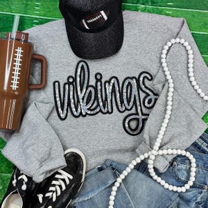 May include: A gray sweatshirt with the word "Vikings" in silver sequins, a black baseball cap with a football design, a brown tumbler, black sneakers, a white beaded necklace, and denim jeans are arranged on a green football field background.