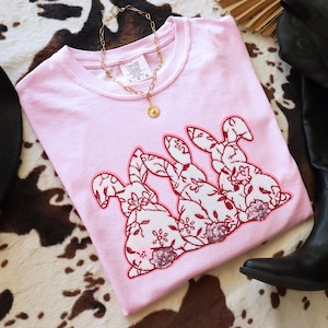 May include: A light pink Comfort Colors sweatshirt featuring three embroidered bunnies with floral patterns in red and white. The bunnies have sequin accents. A gold necklace with a pendant rests on the shirt.