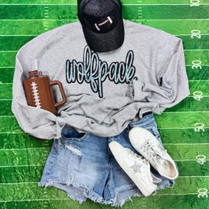May include: A casual outfit featuring a gray sweatshirt with "wolfpack" text, denim shorts, white sneakers, and a black baseball cap with a football design. A brown football-themed tumbler is also included.