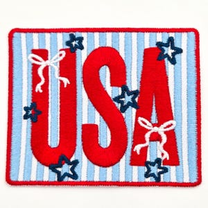 May include: Embroidered patch featuring the word "USA" in bold red letters, adorned with white bows and navy blue stars. The background has vertical stripes of light blue and white, all framed by a red border.
