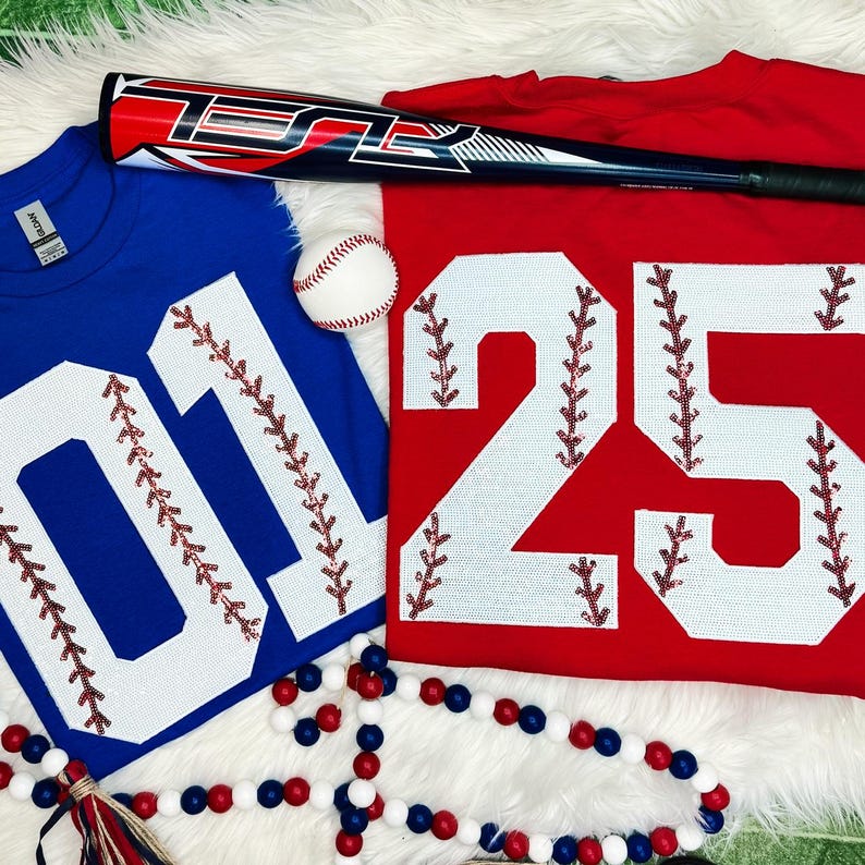 Baseball Number Sequin Patch, close-up of red and blue jerseys with white sequined numbers decorated with red thread stitches and a baseball, baseball bat, and red, white, and blue beads on a white fur background.