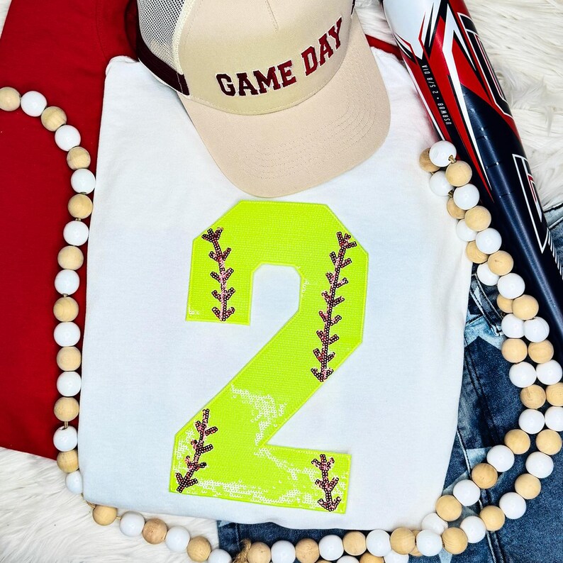 Softball Number Sequin Patch, bright yellow numeral 2 with pink and black embroidered details on a white fabric background, used as a decorative patch for sports apparel.