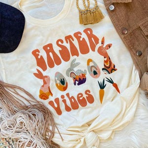 May include: Cream-colored t-shirt with the words "Easter Vibes" in orange, along with Easter-themed graphics. The design includes orange bunnies, decorated eggs, and carrots. The shirt is styled with a corduroy jacket and accessories.
