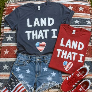 May include: A dark gray t-shirt and a red t-shirt, both featuring white fuzzy lettering that reads LAND THAT I and a heart-shaped American flag patch. Also included are blue denim shorts with white star prints and a pair of red sneakers.