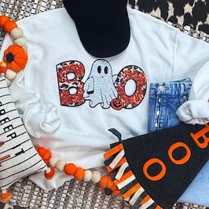 May include: White Halloween-themed sweatshirt with the word "BOO" in red leopard print and a glittery ghost graphic. A black baseball cap and denim jeans are also visible. Decorative orange and white felt ball garland and a black banner with orange letters complete the festive scene.