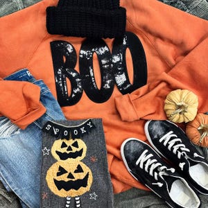 May include: An orange Halloween-themed sweatshirt with the word "BOO" in black sequins, a black knit beanie, and a gray shirt with a pumpkin design. Also included are blue jeans, black sequin sneakers, and decorative pumpkins.