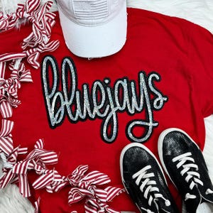 May include: A red t-shirt with the word "Bluejays" in sequined lettering, a white baseball cap with an American flag patch, and black sneakers with white laces. Decorative red and white striped bows are arranged around the shirt.