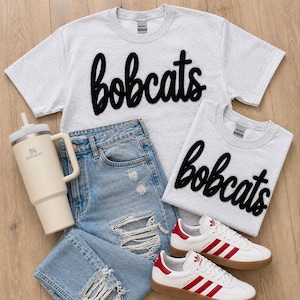 May include: A flat lay featuring a light gray t-shirt and sweatshirt with the word "bobcats" in black script, light blue distressed jeans, a cream-colored tumbler, and white Adidas Gazelle sneakers with red stripes.