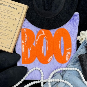 May include: A Halloween-themed flat lay featuring a lavender sweatshirt with the word "BOO" in orange sequins. A black hat, denim jeans, black shoes, and a white beaded necklace are also present. A vintage-style book with text is visible.