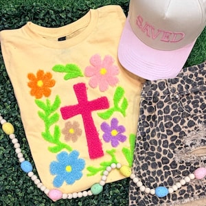 May include: A light yellow t-shirt with a pink cross and colorful floral embroidery. A pink baseball cap with "SAVED" embroidered on it and leopard print denim shorts are also in the image. A beaded garland with pastel-colored egg accents is also present.