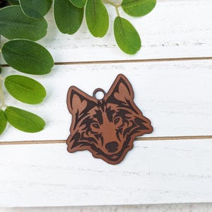 May include: A brown leather wolf pendant with intricate tribal-style detailing. The pendant features a circular hole at the top for hanging. The wolf's face is the focal point, with detailed eyes and fur patterns. The pendant is set against a white wooden background.