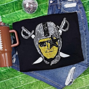 May include: Black t-shirt featuring a sequined design of a football helmet and crossed swords. The helmet is silver and black, with a gold face and a black star. The shirt is paired with denim shorts and a brown tumbler.