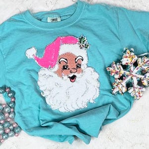May include: A turquoise t-shirt featuring a sequined Santa Claus design with a pink hat and white beard. A decorative snowflake and beaded garland are also visible, suggesting a Christmas theme.