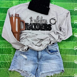 May include: A gray sweatshirt with the text "Touchdown Raiders" in black and silver glitter, paired with denim shorts, a black beanie, and a football-themed tumbler. The tumbler is brown with white stitching details.