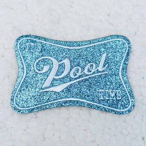 May include: A teal, glittery patch with white text that reads "It's Pool Time". The patch has a unique shape with a white border. The patch is likely for a bag, jacket, or other apparel.
