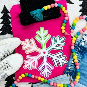 May include: A bright pink long-sleeve shirt with a large, colorful snowflake applique. The image also features a black beanie with a teal tassel, white sneakers, denim jeans, and a beaded garland with rainbow-colored beads.