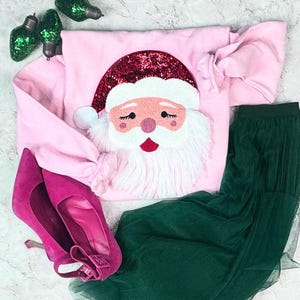 May include: A festive holiday ensemble featuring a pink Santa Claus sweatshirt with a sequined hat and fluffy white beard. Paired with a green tulle skirt and bright pink high heels. Decorative green Christmas light ornaments are also included.