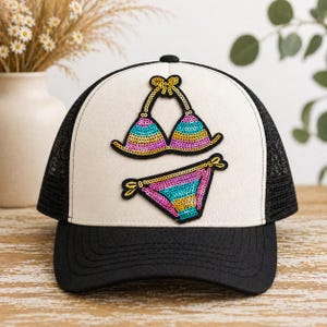 May include: Black and white trucker hat with a white front panel featuring a sequined bikini design. The bikini top and bottom are decorated with colorful sequins in shades of blue, yellow, and pink. The hat has a curved brim and a mesh back.
