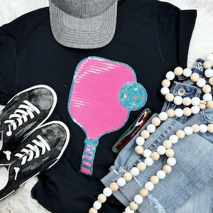 May include: Black t-shirt with a large pink sequined pickleball paddle and a blue sequined ball. The image also includes a gray baseball cap, black sneakers, denim shorts, and a wooden bead garland.