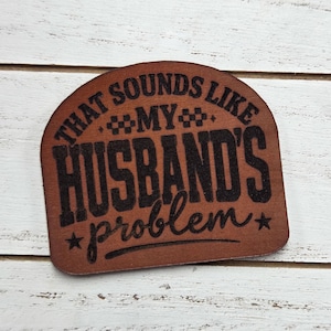 May include: A brown, semi-circular wooden plaque with the phrase "THAT SOUNDS LIKE MY HUSBAND'S problem" in black lettering. The plaque has a rustic, engraved appearance, with stars and decorative elements.