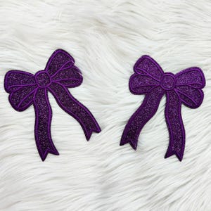 May include: Two purple glitter bow appliques. Each bow has a detailed design with a central knot and long, flowing ribbons. The bows are a vibrant shade of purple, perfect for crafting or embellishing projects.