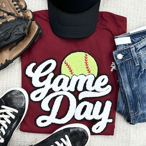 May include: A maroon t-shirt with the words "Game Day" in white script, featuring a yellow softball graphic. The image includes a black baseball cap, blue jeans, black sneakers, and a brown baseball glove.