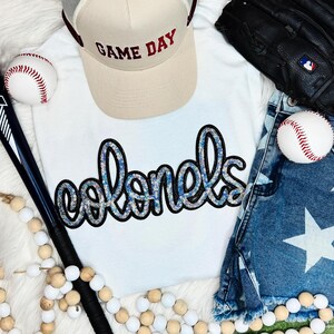 May include: A white t-shirt with the word "colonels" in blue sequins, a beige "Game Day" baseball cap, a baseball, a baseball glove, and denim shorts with a star pattern. A baseball bat and wooden beads are also present.