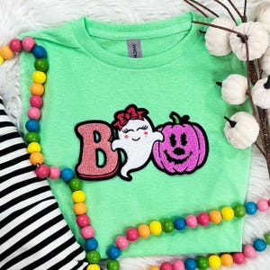 May include: A light green t-shirt with a Halloween-themed design. The design features the word "Boo" with a pink letter "B", a cute ghost with a red bow, and a pink pumpkin. Colorful wooden beads and white pumpkins decorate the background.