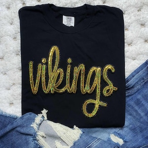 May include: Black t-shirt with the word "Vikings" in gold sequin lettering. The shirt is folded and placed on a white surface with a pair of blue jeans.