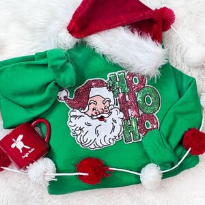 May include: A green Christmas-themed sweatshirt with a Santa Claus design and the words "HO HO HO". The sweatshirt is paired with a red Santa hat with white trim and a red mug with the words "Jingle Bells".