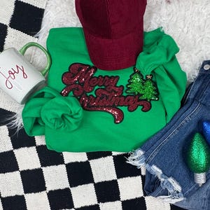 May include: A green sweatshirt with the text "Merry Christmas" in red sequins and two green sequined Christmas trees. A maroon corduroy baseball cap and denim shorts are also in the image. A white mug with the word "Joy" is also visible.