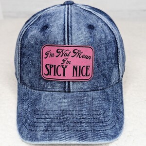 May include: A blue denim baseball cap with a pink rectangular patch. The patch reads "I'm Not Mean I'm SPICY NICE" in a black font. The cap has a curved brim and a button on top.
