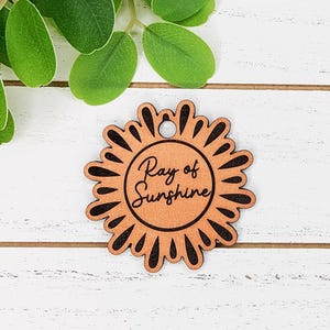 May include: A wooden sun-shaped tag with the words "Ray of Sunshine" in a circular design. The tag has a natural wood color with black accents and a small hole at the top for hanging. The tag is a decorative item.