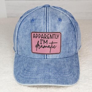May include: A light blue denim baseball cap with a pink patch that reads "Apparently I'm dramatic" in black and pink lettering. The cap has a curved brim and a button on top.