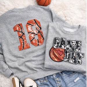 May include: Two gray sweatshirts with basketball-themed designs. One features the number "18" in orange and black, resembling a basketball. The other says "GAME DAY" with a basketball graphic and a heart. A basketball and denim shorts are also in the image.