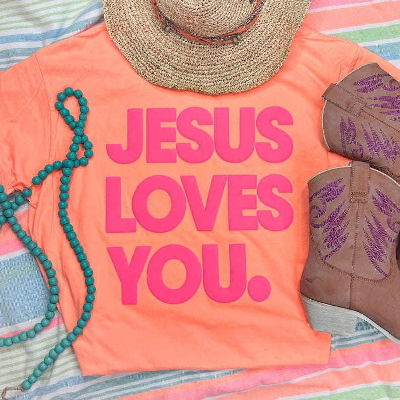 Product Jesus Loves You Pink PUFF Transfer, pink t-shirt with bold lettering, cowboy boots with purple wing designs, turquoise beaded necklace, and a large straw hat on a colorful striped fabric background.