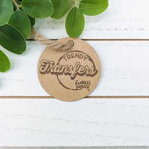 May include: A round, brown paper tag with the words "Trendy Transfers" and "Express Yourself" printed in a dark brown color. The tag is attached to a piece of twine and is set against a white wooden background with green leaves.