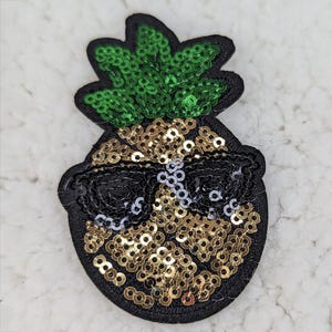 May include: A sequined pineapple patch with black sunglasses. The pineapple body is covered in gold sequins, with a black outline. The top has green sequins. This decorative patch can be sewn or ironed onto clothing or accessories.